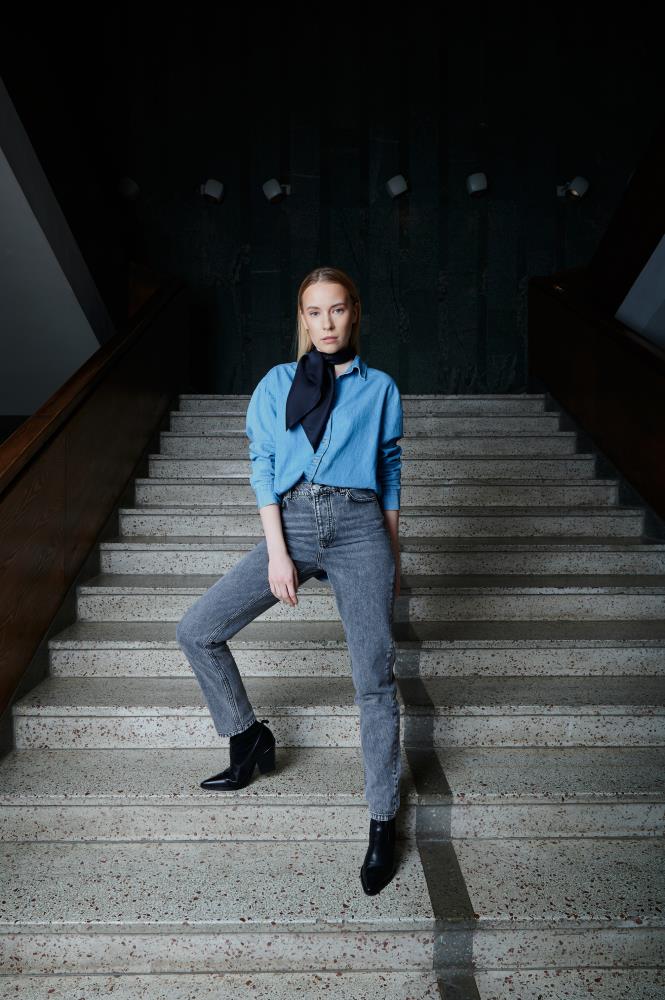 A person stands confidently on a staircase, wearing a light blue shirt, dark midnight blue silk scarf, gray jeans, and black boots. The setting is dimly lit and stylish.