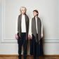 Two models, one male and one female, wearing beige and brown wool scarves of the SKARELE brand, standing against a plain background.