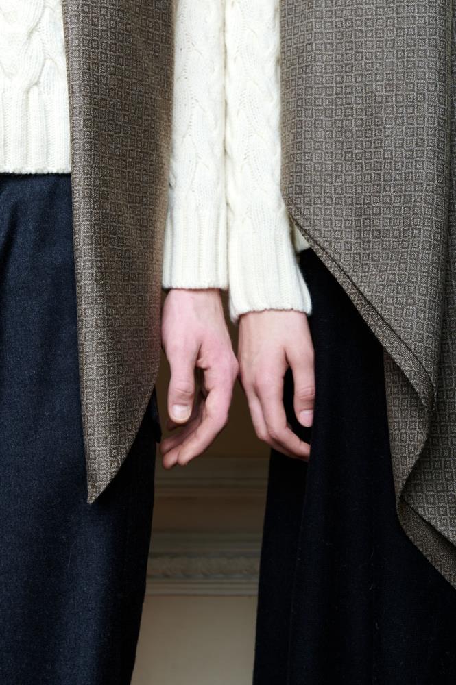A close-up photo of two figures standing side by side, showcasing clothing details. Both individuals wear light-colored cable-knit sweaters and dark trousers, with a brown scarf made of high-quality wool and featuring a fine geometric pattern draped over their shoulders. In the center, two hands are positioned close together, creating a sense of closeness. The composition highlights the texture of the wool fabric, its coziness, and the color harmony.