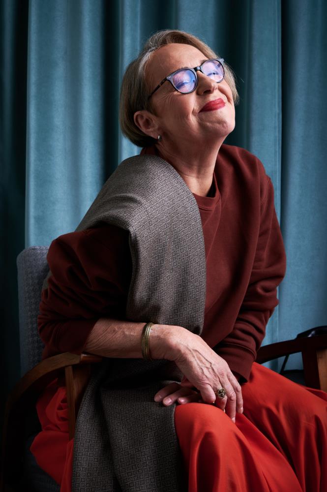 A smiling woman with short hair and glasses sits in an armchair against a backdrop of blue curtains. She is wearing a burgundy sweatshirt, with a greyish -brown-beige wool scarf featuring a fine geometric pattern stylishly draped over her shoulder. The woman has her head tilted slightly upward, one hand resting on the other, and the overall atmosphere of the photo radiates coziness, elegance, and inner peace.
