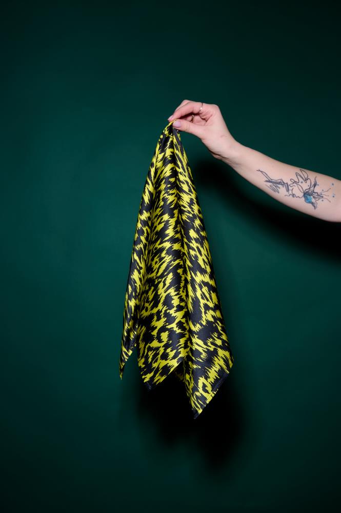 A hand with a tattoo holding a vibrant yellow and black patterned silk scarf against a dark green background. The photo highlights the fabric's sheen, dynamic graphic pattern, and high-quality texture. A stylish and contrasting accessory that adds a modern accent 