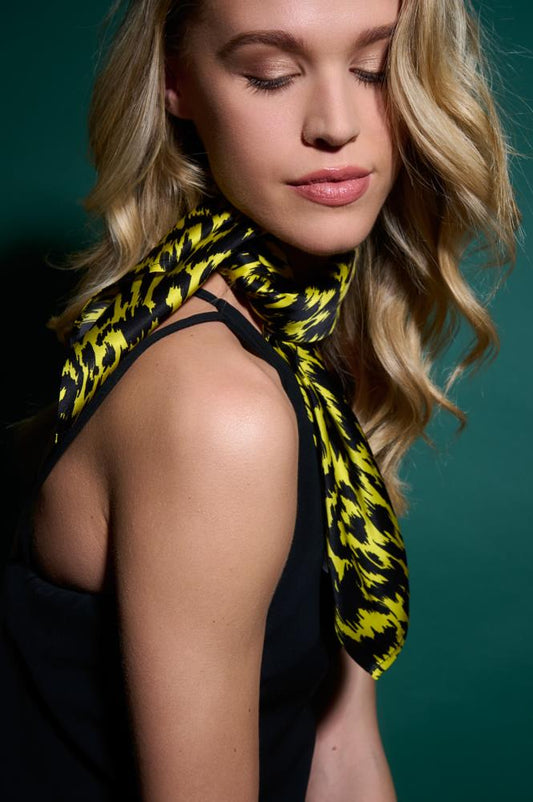 A model portrait showcasing a stylish yellow and black patterned silk scarf tied around her neck. The bold graphic pattern and soft fabric sheen stand out contrastingly against a dark top and a deep green background