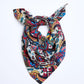 A colorful patterned scarf tied into a knot and laid against a clean white background. The accessory features a rich paisley and botanical pattern, combining vibrant blue, red, yellow, green, and black colors. The scarf is arranged in a triangular shape, highlighting the fabric's softness and the intensity of the colors.