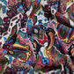 A close-up of crumpled fabric with an extremely rich and colorful pattern. The surface of the fabric features a blend of classic paisley, floral, and geometric motifs. The color palette is intense, dominated by vibrant blue, red, yellow, green, and black on a white background. The folds of the material create a sense of depth and highlight the intricacy of the pattern.