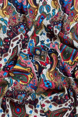 A close-up of crumpled fabric with an extremely rich and colorful pattern. The surface of the fabric features a blend of classic paisley, floral, and geometric motifs. The color palette is intense, dominated by vibrant blue, red, yellow, green, and black on a white background. The folds of the material create a sense of depth and highlight the intricacy of the pattern.