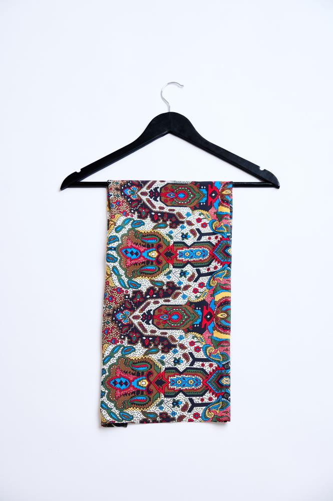 A folded scarf hanging on a black wooden hanger against a white background. The accessory features a very bright and dense pattern combining paisley, floral, and ethnic motifs. The color scheme is dominated by rich red, blue, yellow, and green, creating a contrasting and vibrant look. The scarf hangs straight, highlighting the symmetry of the pattern and the quality of the fabric.