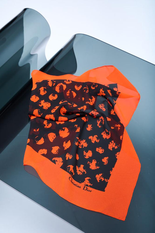 A silk scarf with orange edges and a black and orange pattern, displayed on a reflective surface.