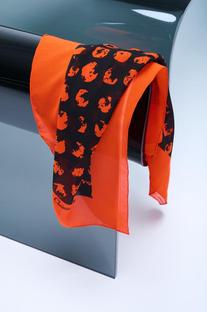 A luxury silk scarf in vibrant orange and black, featuring a bold abstract leopard-style print, elegantly draped over a modern curved glass chair.