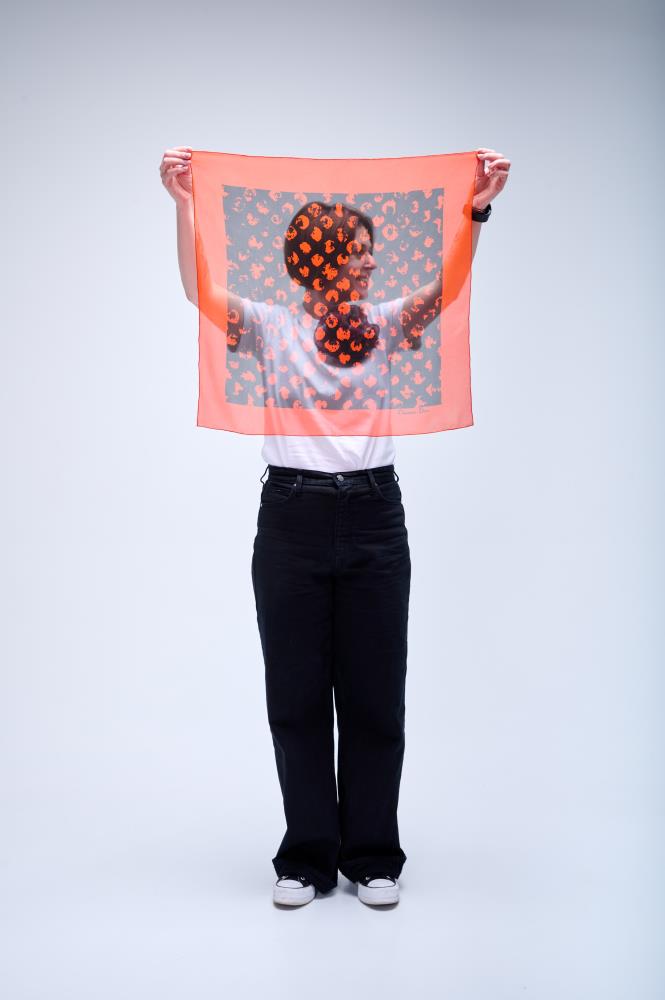 A person holding up a square, semi-transparent  silk scarf in vibrant orange, featuring a dark center panel with an abstract orange leopard-style print.