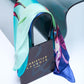 Vibrant Christian Lacroix silk scarf with a colorful floral print in turquoise, purple, and green, elegantly draped over a glass surface next to its luxury designer gift box.