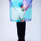 A person holding up a large, translucent Christian Lacroix silk scarf featuring a vibrant purple and pink floral motif on a gradient turquoise and blue background.