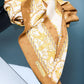 Luxury silk scarf with a baroque-style print featuring gold chains, mythical griffins, and ornate  in mustard yellow and cream white, draped on a dark glass surface.