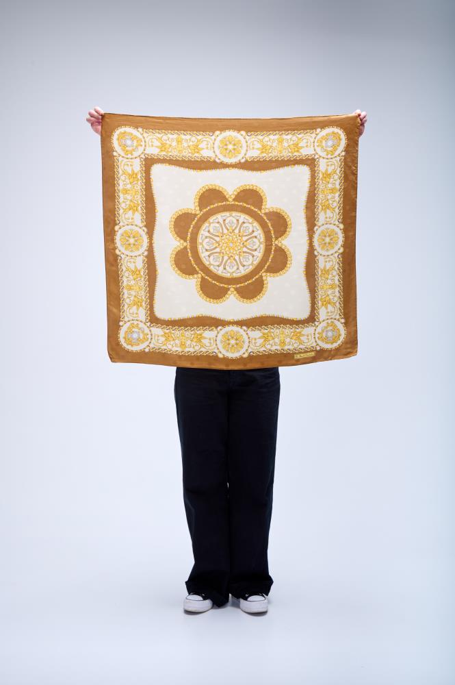 A person holding up a large square silk scarf with a classical baroque design, featuring a central medallion, gold chains, and ornate mustard yellow borders on a white background