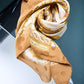 Close-up of a silk scarf artfully swirled on a reflective surface, featuring a mustard gold chain and polka dot pattern with classical baroque ornaments.