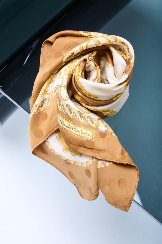 Close-up of a silk scarf artfully swirled on a reflective surface, featuring a mustard gold chain and polka dot pattern with classical baroque ornaments.