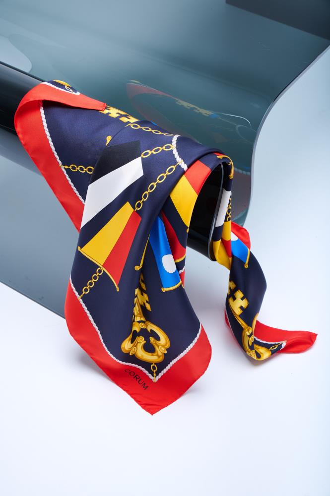 silk scarf with a nautical design featuring gold keys, chains, and maritime signal flags, elegantly draped over a curved glass surface in a minimalist studio.