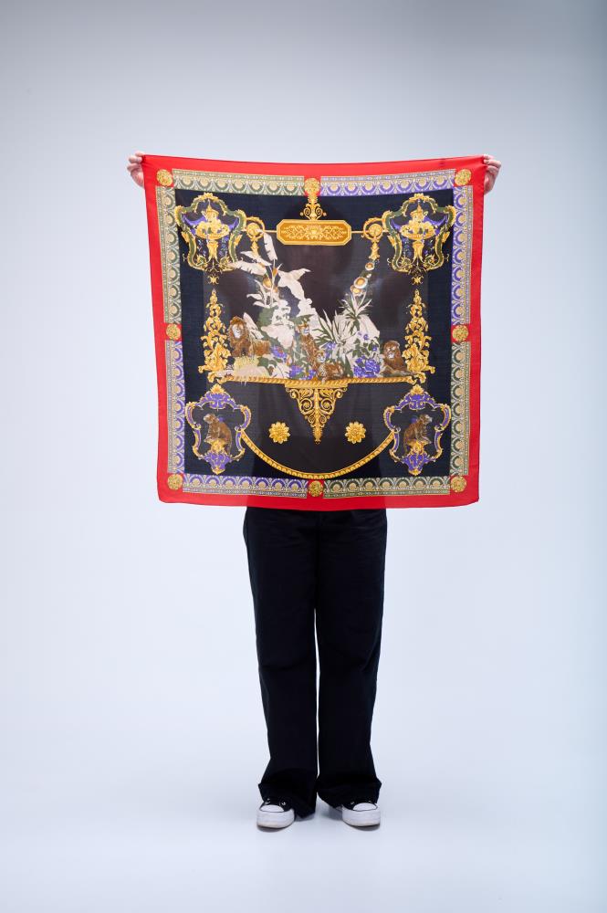 A person holding up a large square silk scarf with a baroque-style design, featuring lions, tropical birds, and ornate gold motifs on a black and red background.