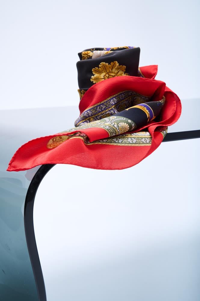 luxury silk scarf with a baroque-style print featuring gold ornaments and a vibrant red border, elegantly draped over a curved modern glass surface in a bright studio.