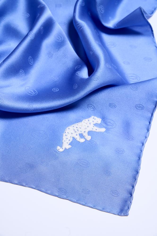 A close-up of a luxuriously draped royal blue silk scarf featuring a subtle monogram pattern and a detailed white panther embroidery on the corner.
