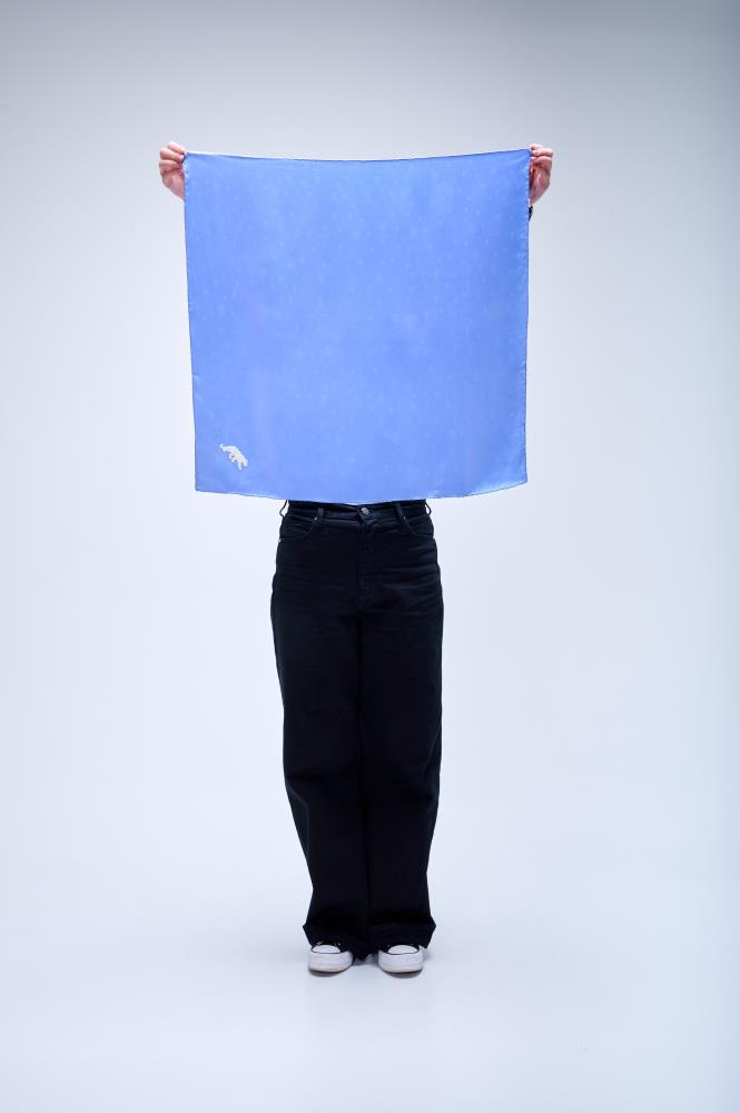 A person holding up a large square royal blue silk scarf with a small white panther embroidery in the corner, standing against a neutral studio background.