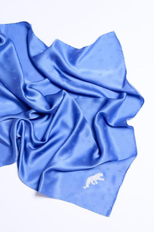 Artfully draped royal blue silk scarf on a white background, featuring a subtle monogram pattern and a distinct white embroidered panther motif in the bottom corner.