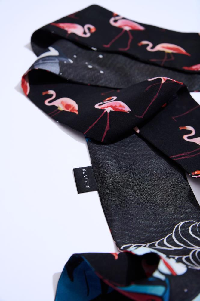A narrow black "Twilly" silk scarf featuring a vibrant pink flamingo pattern and blue accents, folded on a white background. A small black label with the text "SKARELE" is visible. A modern and stylish accessory that adds a playful touch to any look.