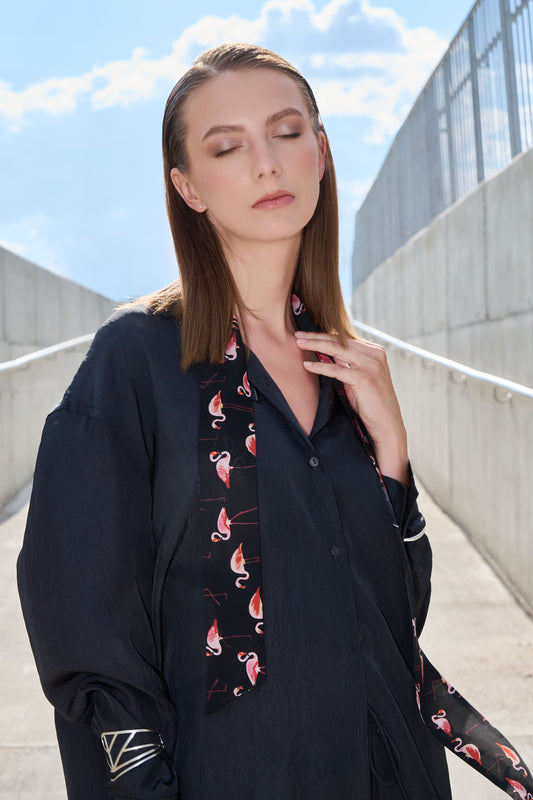 A stylish portrait of a woman in an industrial setting with concrete walls. The model is wearing a loose-fit dark shirt and a narrow "Twilly" silk scarf featuring a vibrant pink flamingo pattern on a black background. The accessory adds a modern, elegant, and playful accent to the look.