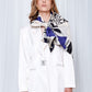 A woman poses confidently in a white blazer with a large patterned scarf in blue and black draped around her neck. Her expression is calm and poised.