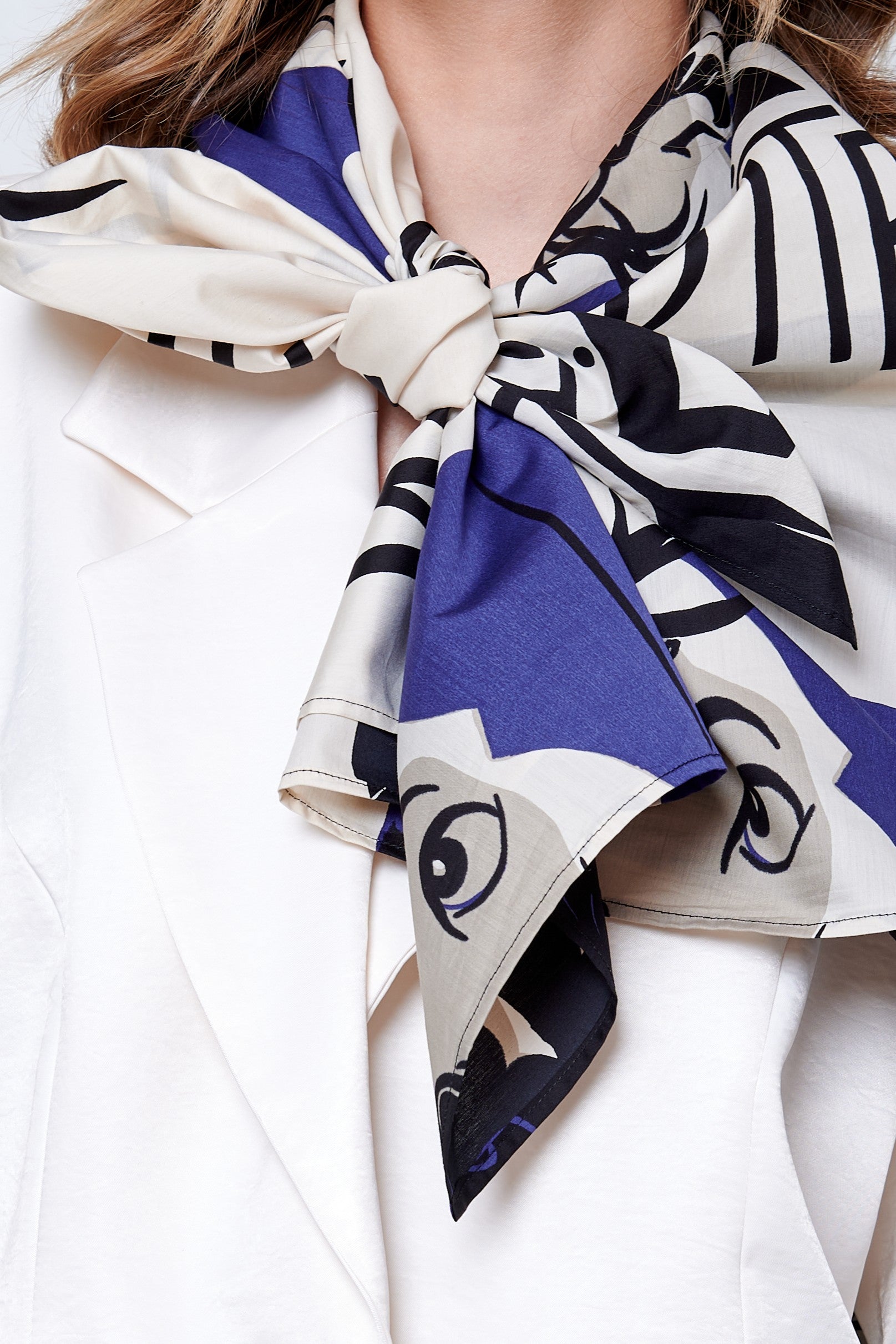 A close-up of a person wearing a white blazer and an abstract patterned scarf in blue, cream, and black, tied in a large, elegant bow at the neck.