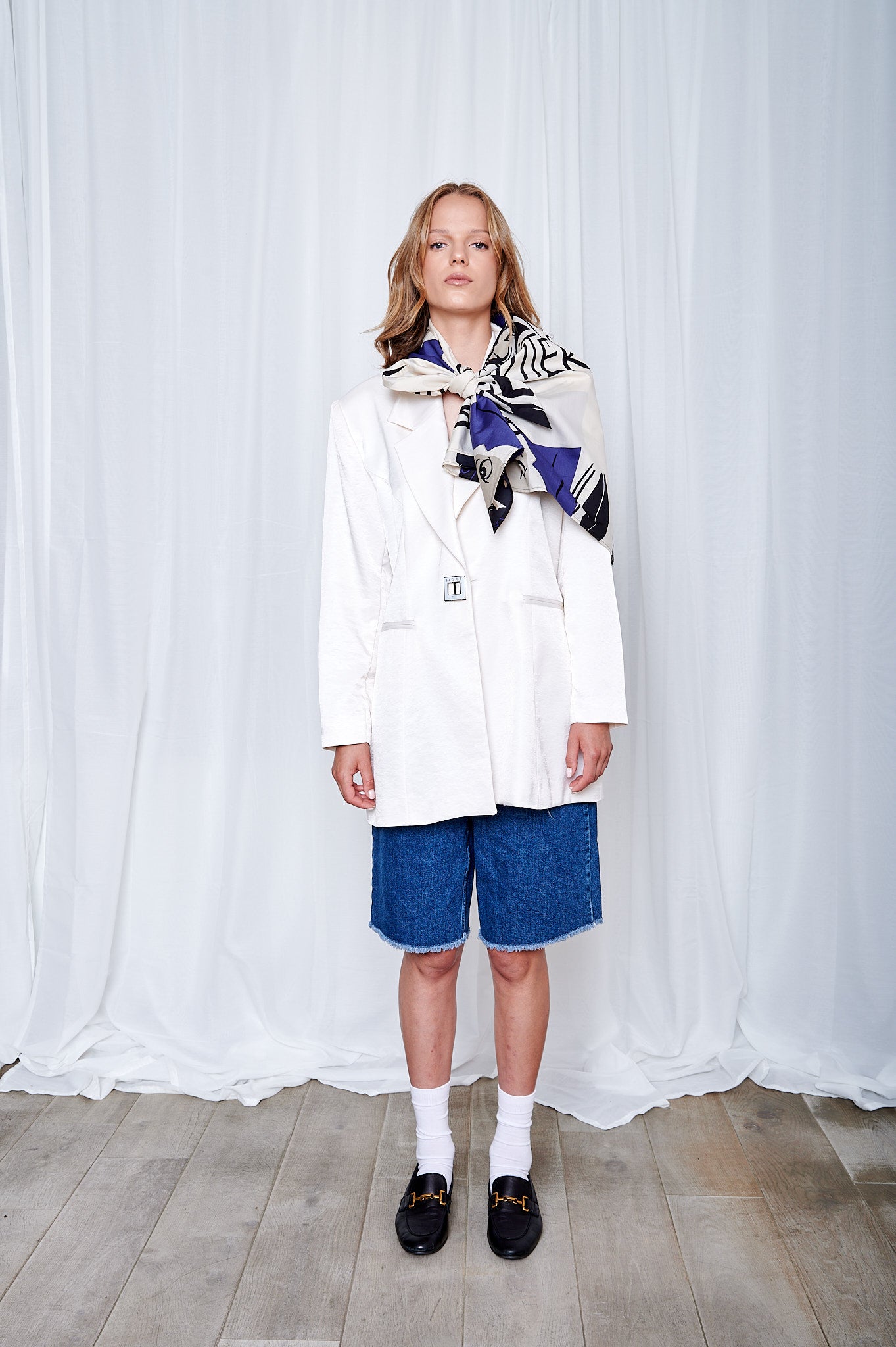 A person stands against a white curtain backdrop wearing a white blazer, denim shorts, and a patterned scarf. The mood is stylish and contemporary.