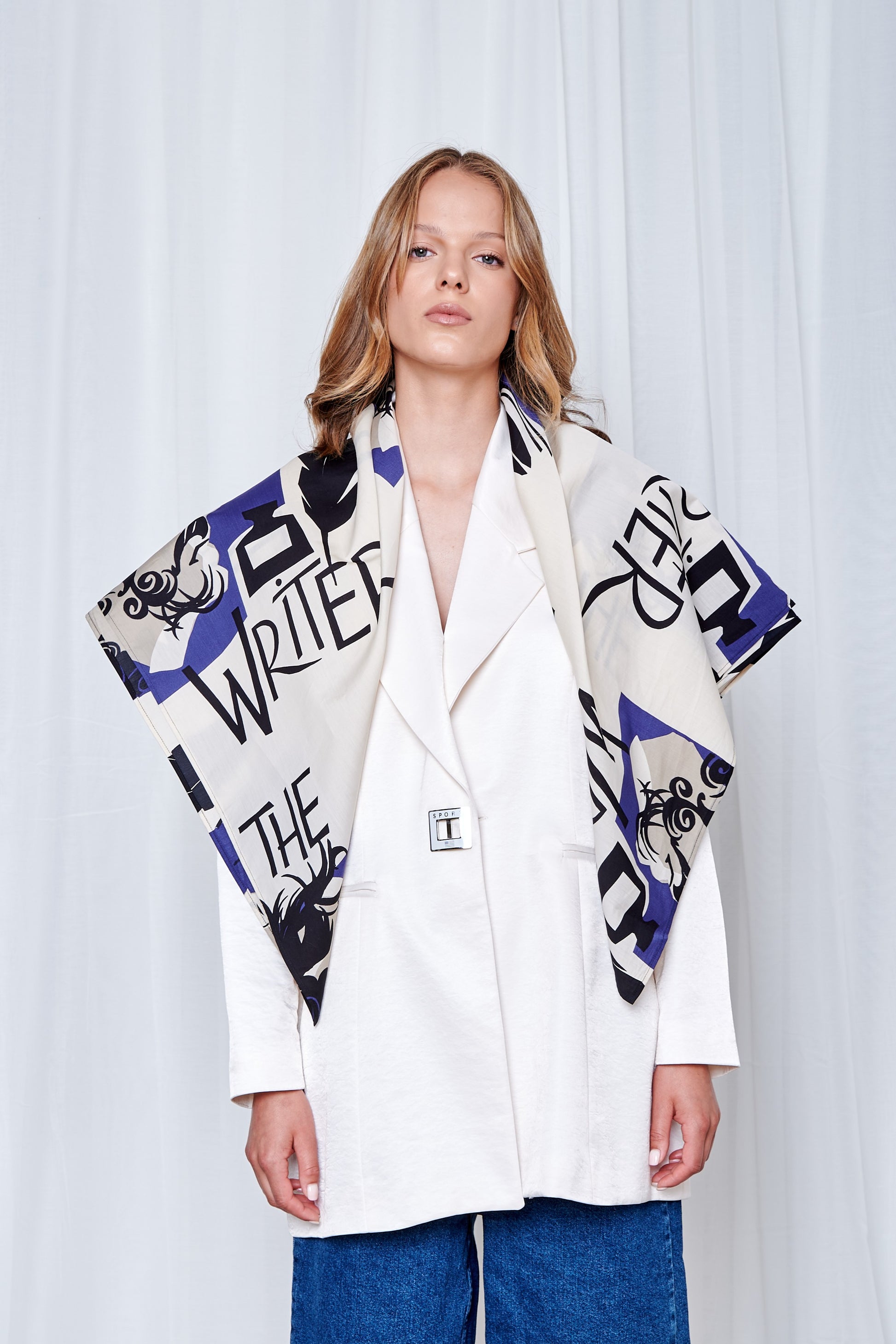 A woman wearing a white blazer and blue jeans stands confidently. A large scarf with bold text and geometric patterns is draped over her shoulders.