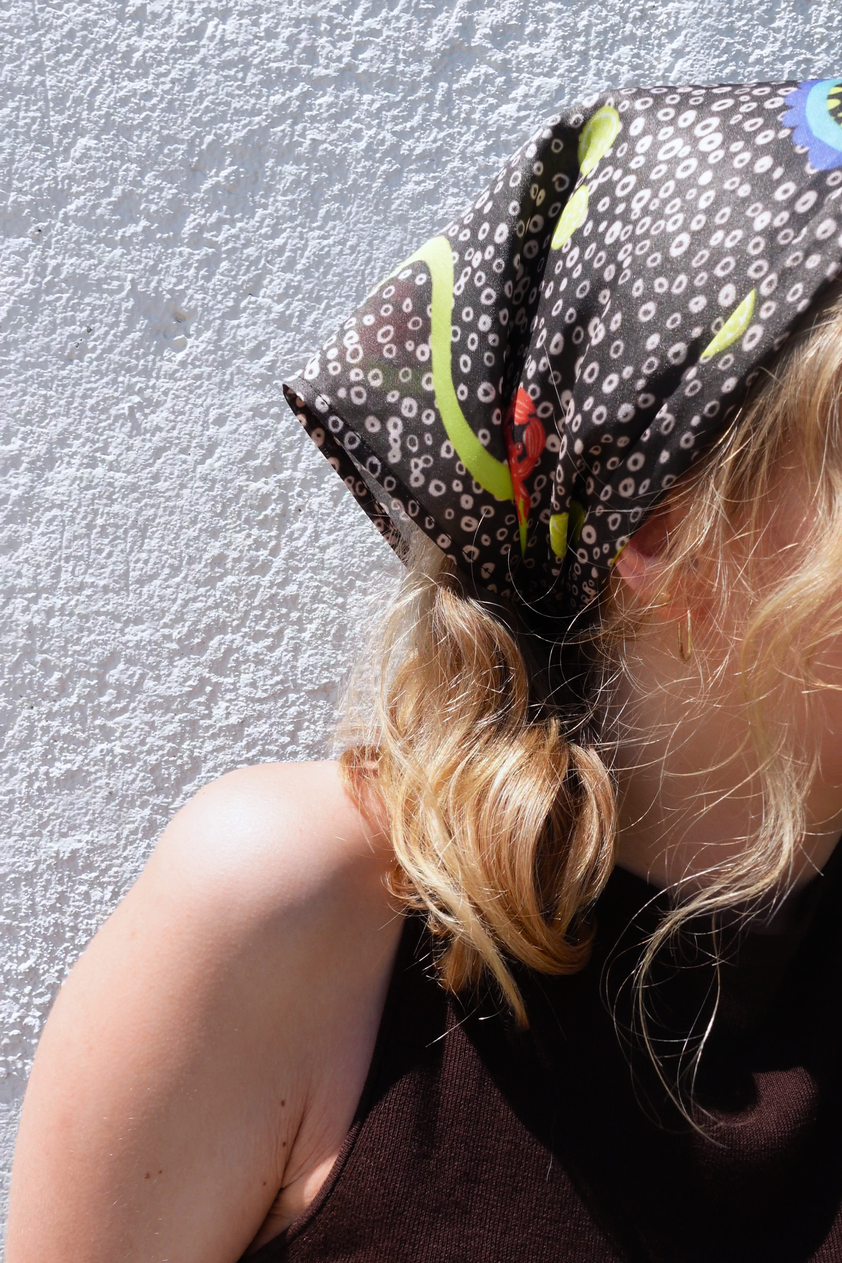 Close-up shot of SKARELE "Summer La Dolce Vita" dark brown silk blend headscarf with vibrant green and blue abstract pattern, tied as a bandana on a blonde woman.