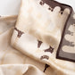 A folded silk scarf with a beige and cream geometric pattern and dark brown abstract motifs, displayed on a light textured background.
