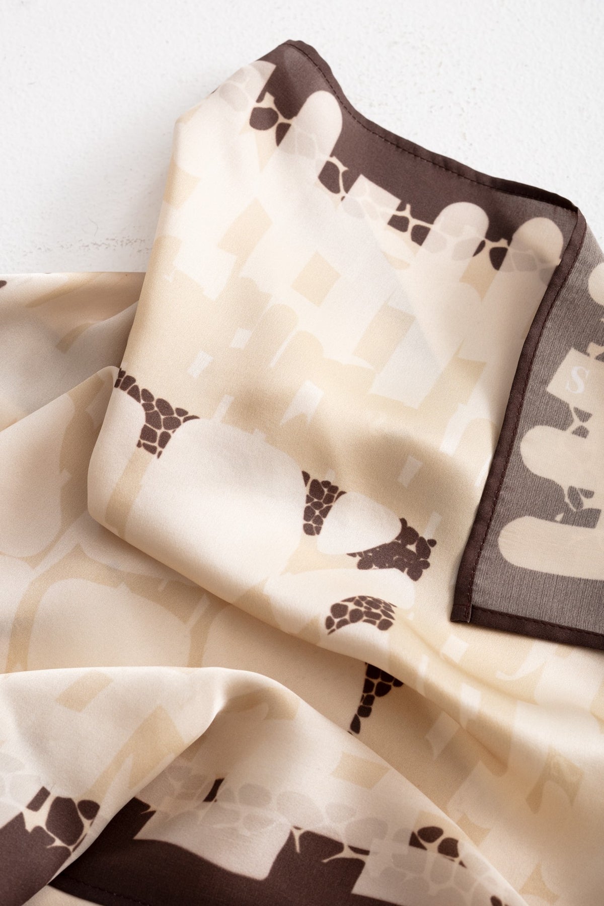 A folded silk scarf with a beige and cream geometric pattern and dark brown abstract motifs, displayed on a light textured background.