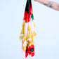 A hand with a distinctive green manicure and a detailed black and blue tattoo is holding a draped silk scarf. The scarf features a bold graphic print with a black and red color block top, large stylized red poppy flowers on a white background, and complex yellow feather-like tassels, set against a light background.