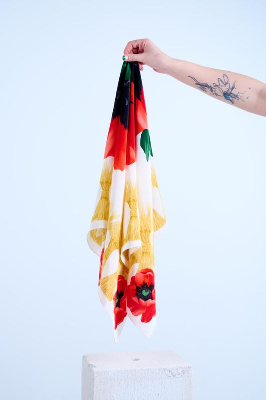 A hand with a distinctive green manicure and a detailed black and blue tattoo is holding a draped silk scarf. The scarf features a bold graphic print with a black and red color block top, large stylized red poppy flowers on a white background, and complex yellow feather-like tassels, set against a light background.