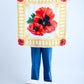 A person in blue jeans holding up a large square Escada silk scarf. The design features a giant red poppy center motif on a white background, surrounded by a yellow decorative tassel border and smaller poppies in each corner.