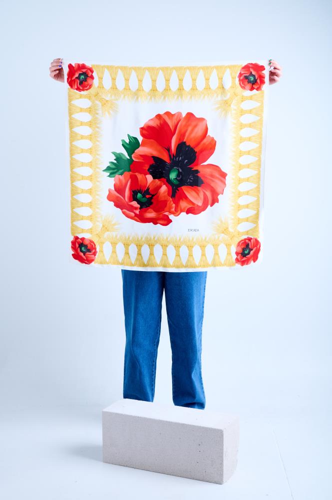 A person in blue jeans holding up a large square Escada silk scarf. The design features a giant red poppy center motif on a white background, surrounded by a yellow decorative tassel border and smaller poppies in each corner.