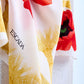 A close-up of a luxury Escada scarf featuring a vibrant red poppy print and intricate yellow wheat-like tassel motifs, elegantly draped over a white stone surface to show the fine fabric texture.