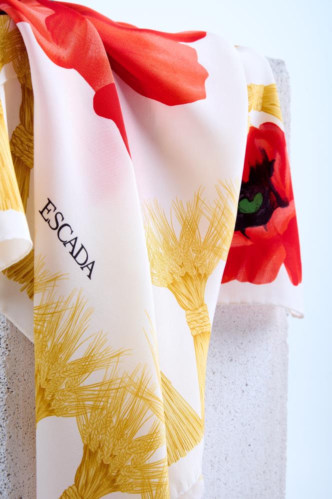 A close-up of a luxury Escada scarf featuring a vibrant red poppy print and intricate yellow wheat-like tassel motifs, elegantly draped over a white stone surface to show the fine fabric texture.