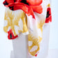 A luxury Escada silk scarf featuring a vibrant red poppy floral print and yellow tassel motifs, artfully draped over a white stone pedestal in a bright studio setting.