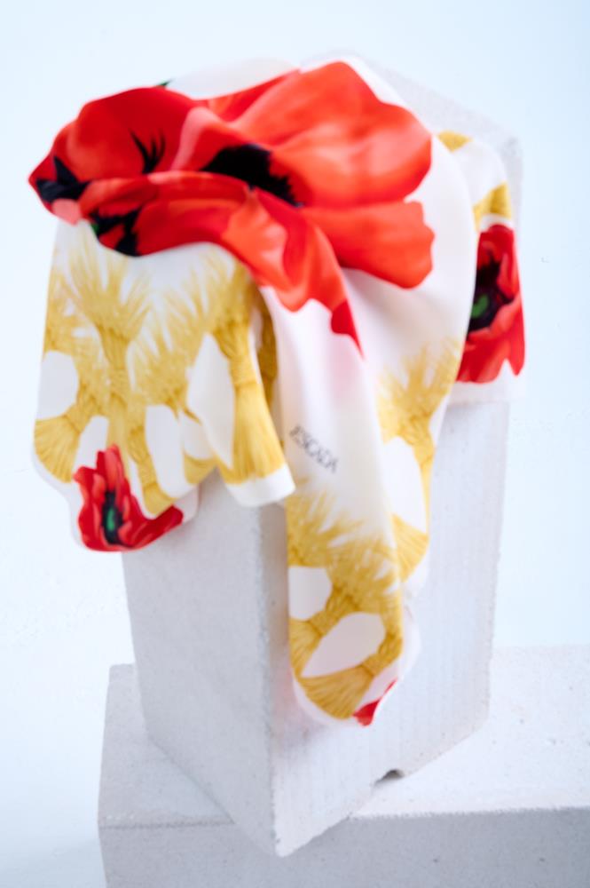 A luxury Escada silk scarf featuring a vibrant red poppy floral print and yellow tassel motifs, artfully draped over a white stone pedestal in a bright studio setting.