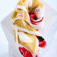 silk scarf artfully swirled into a rose-like shape on a white stone pedestal. The design features vibrant red poppy petals at the center, surrounded by intricate yellow tassel motifs on a clean white background.