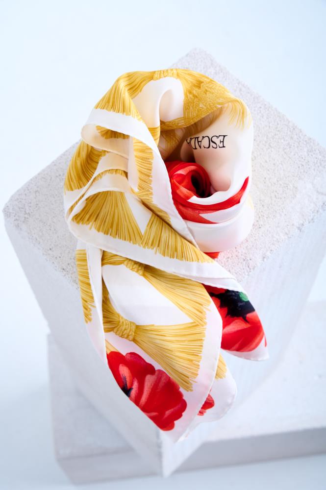 silk scarf artfully swirled into a rose-like shape on a white stone pedestal. The design features vibrant red poppy petals at the center, surrounded by intricate yellow tassel motifs on a clean white background.