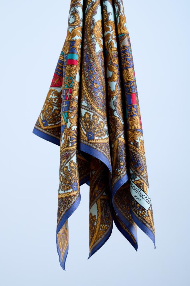A silk scarf featuring an intricate heraldic print with paisley motifs, royal crests, and golden baroque details, elegantly draped to showcase the fluid fabric against a neutral studio background