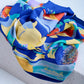 A vintage silk scarf by Piemon d'Urville featuring a vibrant abstract underwater-themed print with yellow, orange, and turquoise motifs, artfully bunched and draped over a white textured stone pedestal in a studio setting.