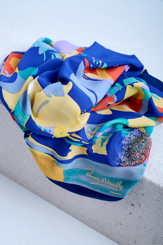 A vintage silk scarf by Piemon d'Urville featuring a vibrant abstract underwater-themed print with yellow, orange, and turquoise motifs, artfully bunched and draped over a white textured stone pedestal in a studio setting.