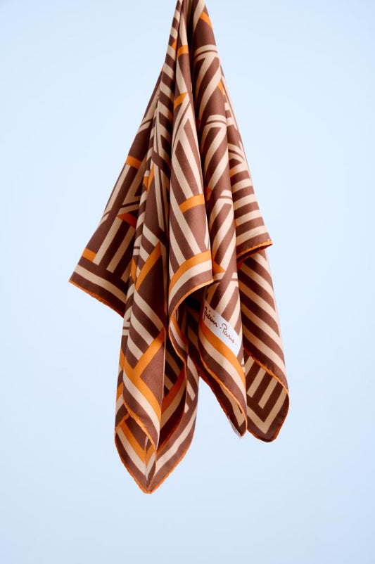 A luxury silk scarf featuring a bold geometric pattern with brown and cream stripes and vibrant orange accents, elegantly draped to showcase the fluid fabric against a neutral studio background.