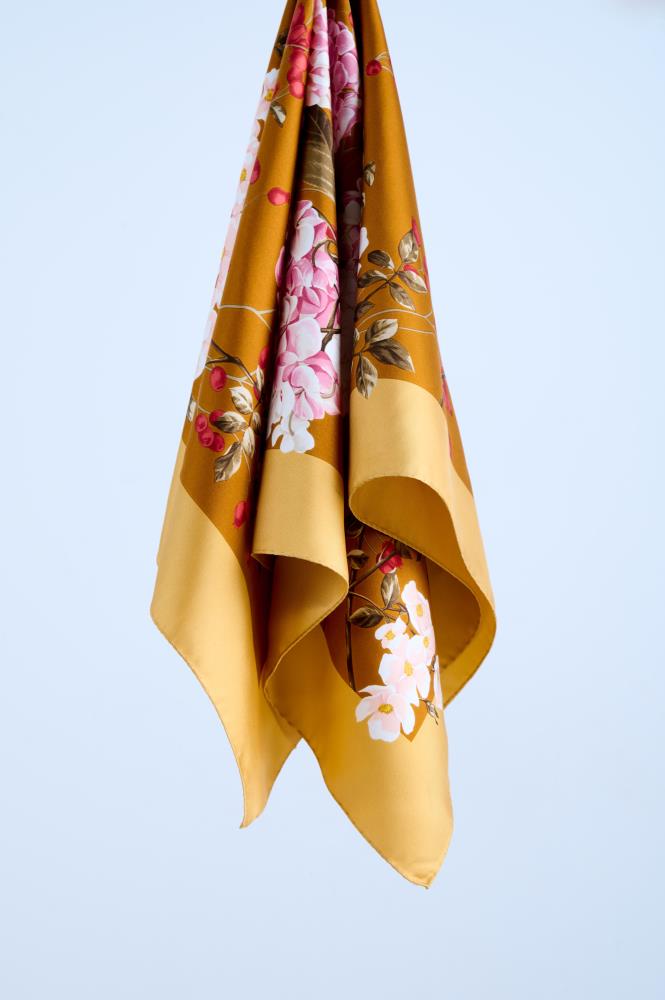 A luxury silk scarf in ochre yellow featuring a vibrant botanical print with pink hydrangeas and white cherry blossoms, elegantly draped to showcase the fluid fabric and rich golden border in a studio setting.