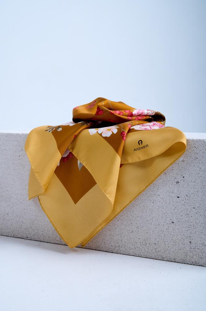 A luxury Aigner silk scarf in a warm ochre yellow hue, featuring a vibrant pink hydrangea and white floral print, artfully draped over a white stone pedestal in a studio setting.